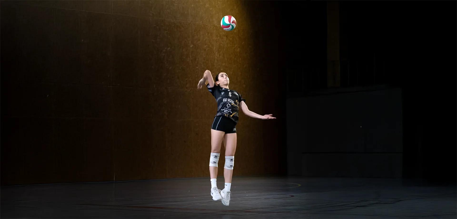 Une joueuse d'une équipe de jeunes au service. Une lumière orangée l'éclaire. Le reste de la salle semble dans la pénombre.