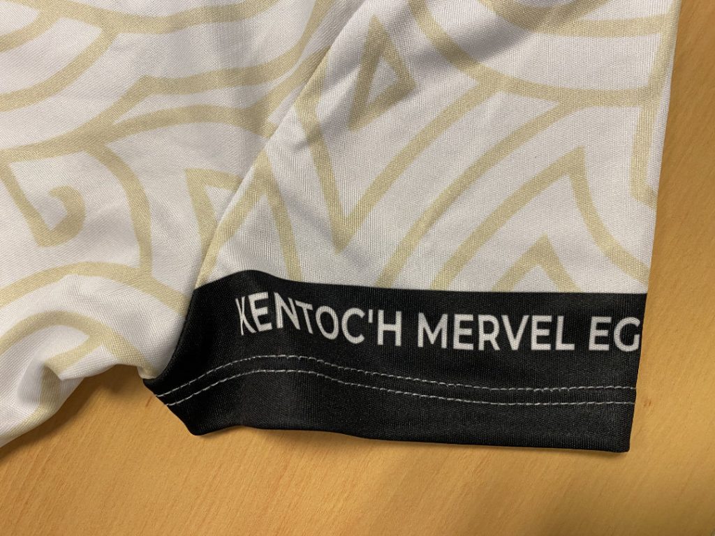 Détail de la manche du maillot extérieur de l'équipe féminine du REC Volley. Le maillot est blanc avec une trame dorée en motif celtique. Le liseré de la manche est noir et il est écrit "Kentoc'h Mervel...", le début de la devise de la Bretagne.